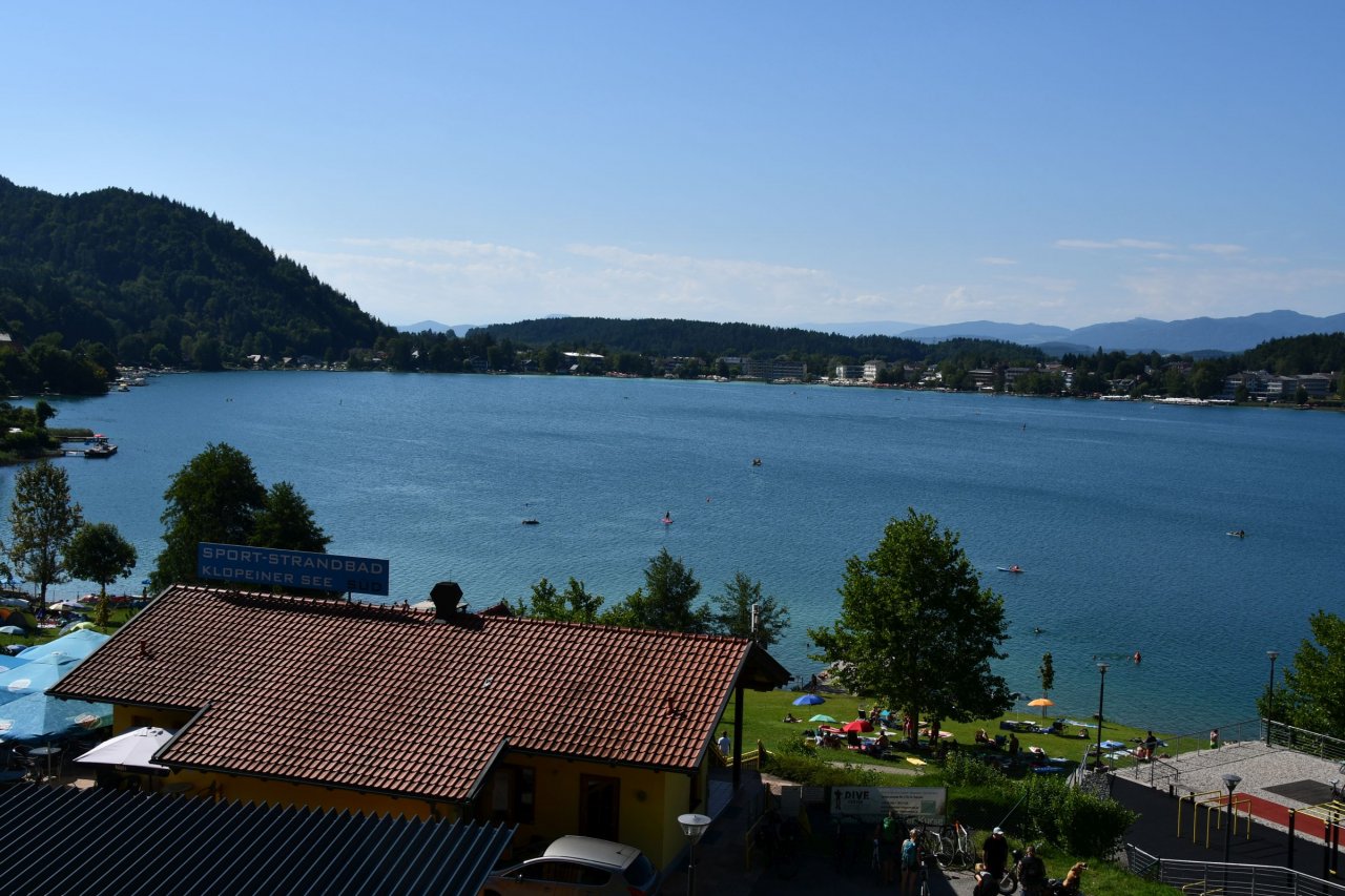 Klopinjsko jezero v svojem sončnem dnevu - turizem cveti, tam nekje na vodi pa je drobcena pika, ki plava na drugo stran.